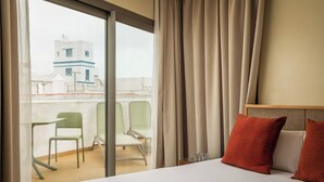 Una habitación de hotel con cama, dos sillas, un balcón con vista a edificios y una cortina.