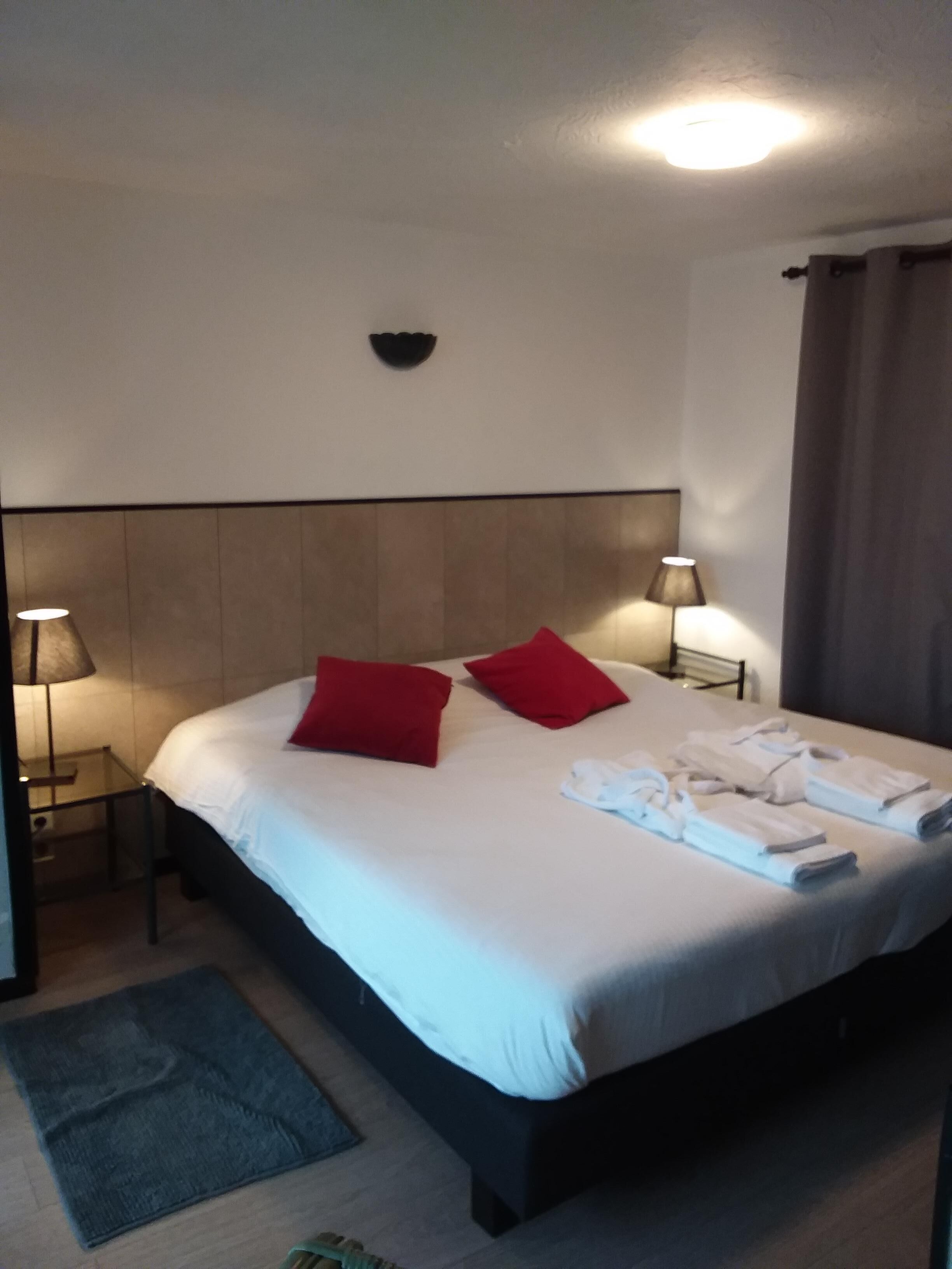 Ein Hotelzimmer mit einem großen Bett, zwei roten Kissen, einem Kopfteil, Nachttischlampen, einem kleinen Teppich und ordentlich gefalteten Handtüchern auf dem Bett.