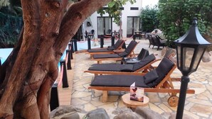 Zona junto a la piscina con sillones de descanso, suelo de piedra y un gran árbol.