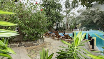 Un área de piscina interior con suelo de piedra, sillones de madera y abundante vegetación.