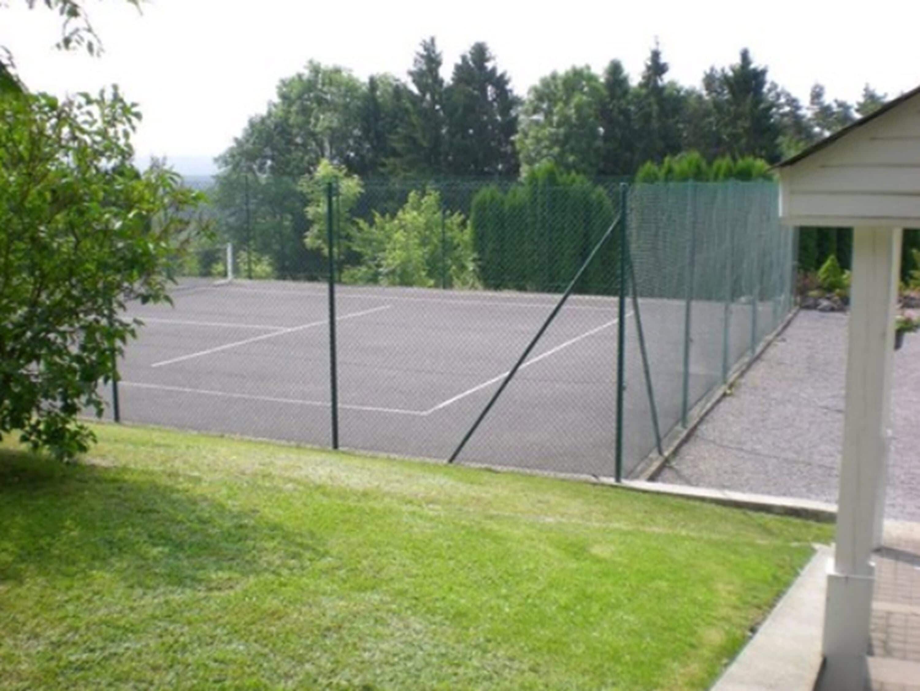 A tennis court with a chain-link fence, surrounded by trees and a building.