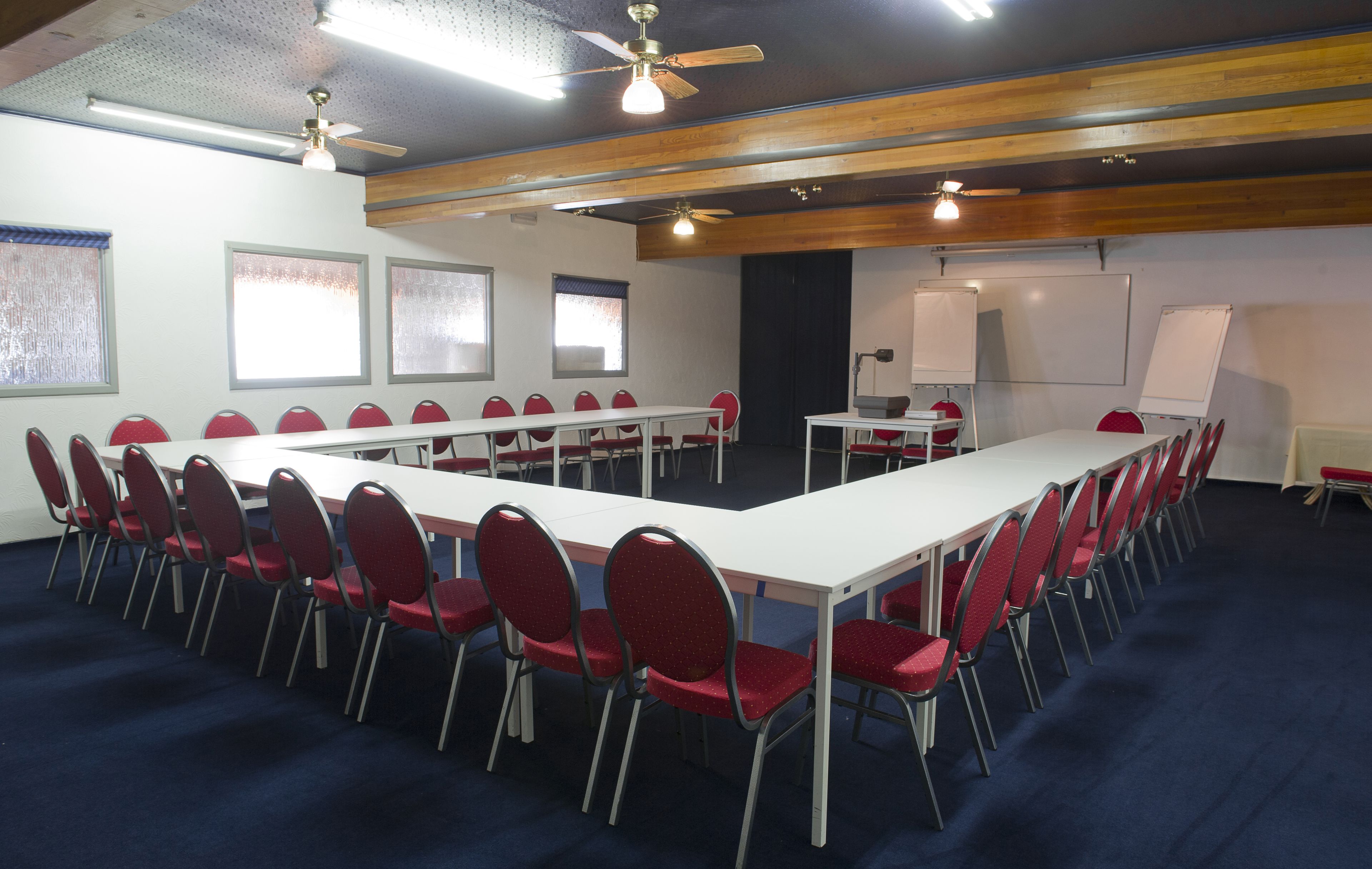 Un salón de conferencias con una larga mesa, sillas rojas, una pizarra y ventiladores de techo.
