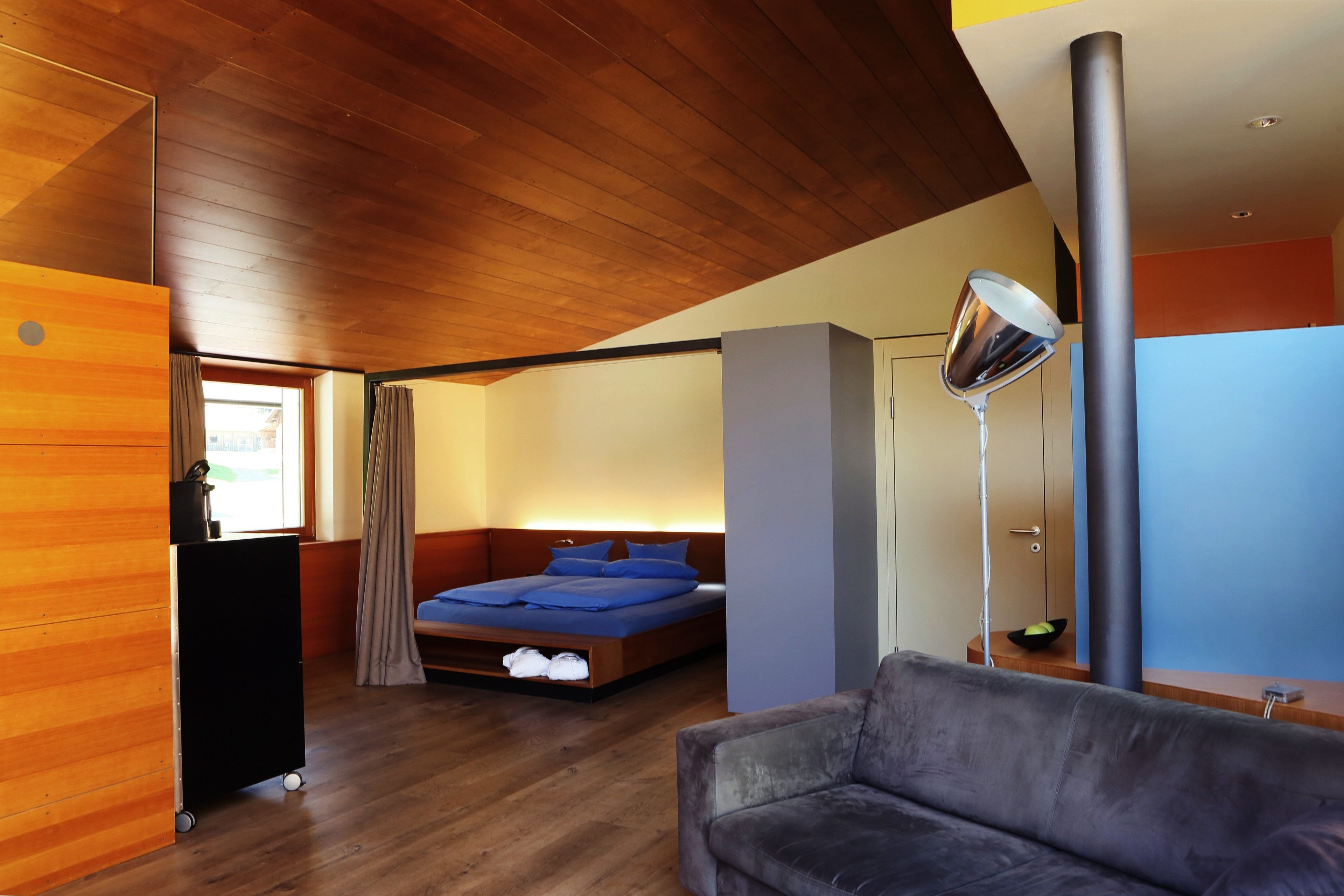 Un dormitorio moderno con techo de madera, cama con ropa de cama azul, sofá gris y un armario negro.