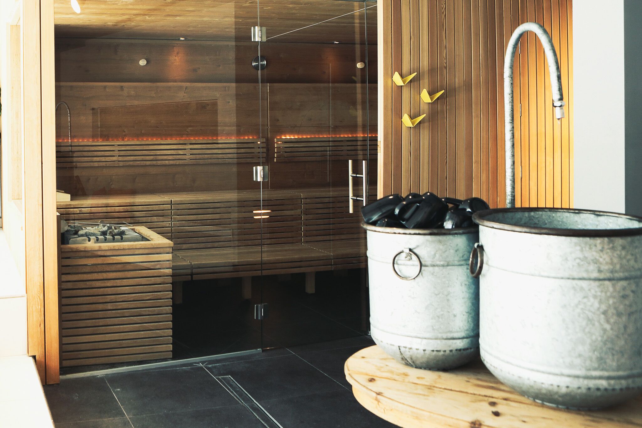 A sauna with wooden walls, a glass door, and a wooden bench. There are two metal pots on a wooden table.