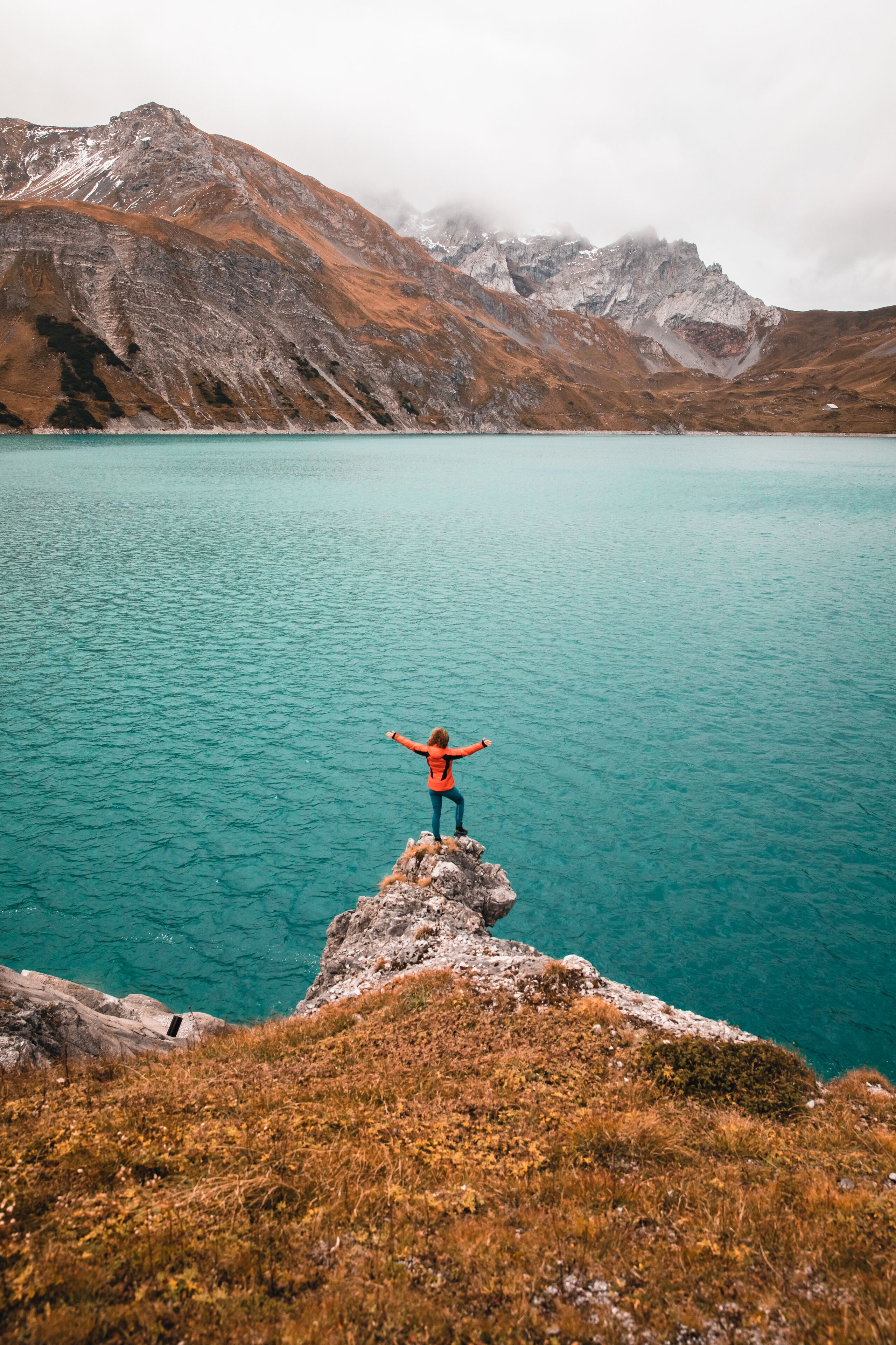 A person standing on a rocky outcrop with arms outstretched, overlooking a turquoise lake and mountains in the background.