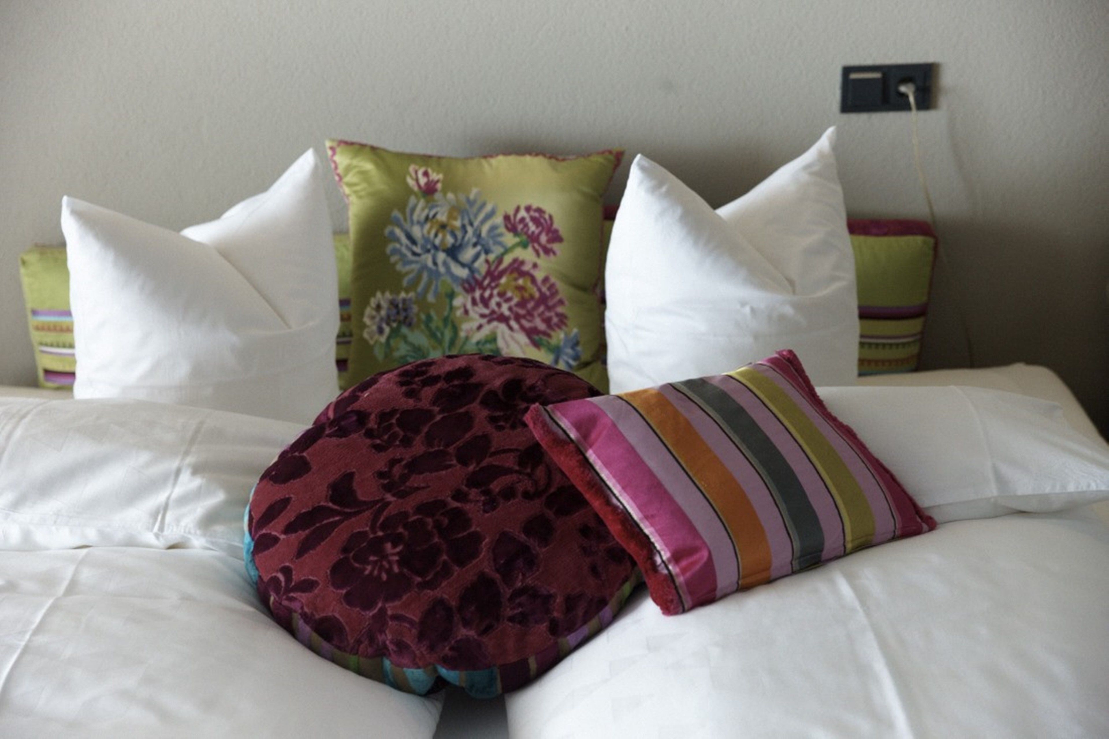 Un lit avec du linge de lit blanc, un coussin à motifs floraux, un coussin à rayures et un coussin à motifs floraux foncés.