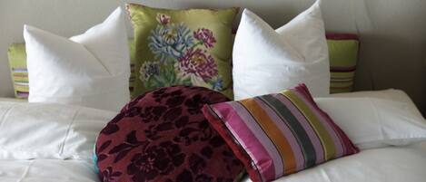 Un lit avec du linge de lit blanc, un coussin à motifs floraux, un coussin à rayures et un coussin à motifs floraux foncés.