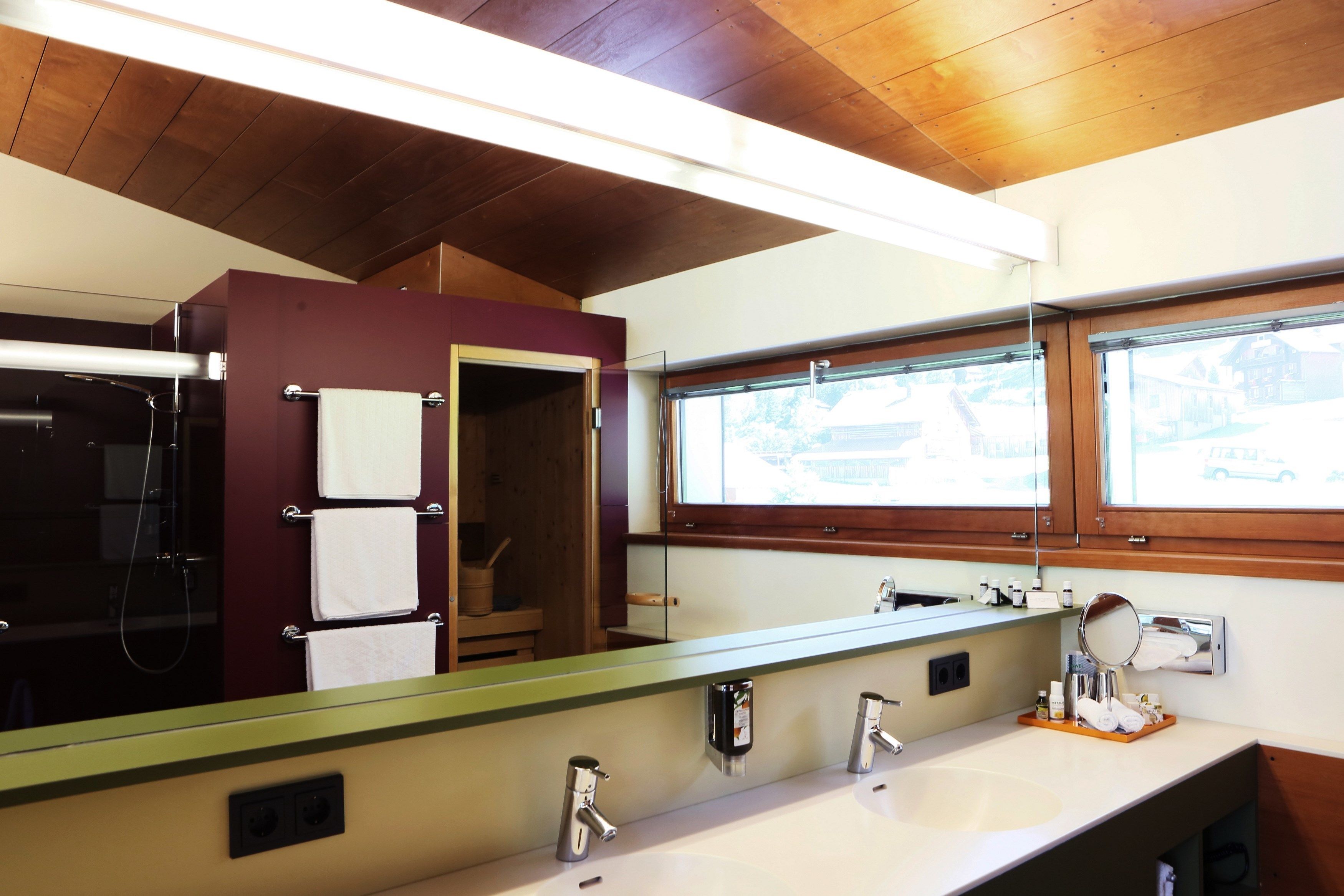 Un bagno con soffitto in legno, uno specchio grande, un lavandino con due rubinetti e una finestra con vista sugli edifici.