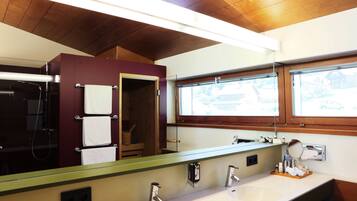 A bathroom with a wooden ceiling, a large mirror, a sink with two faucets, and a window with a view of buildings outside.