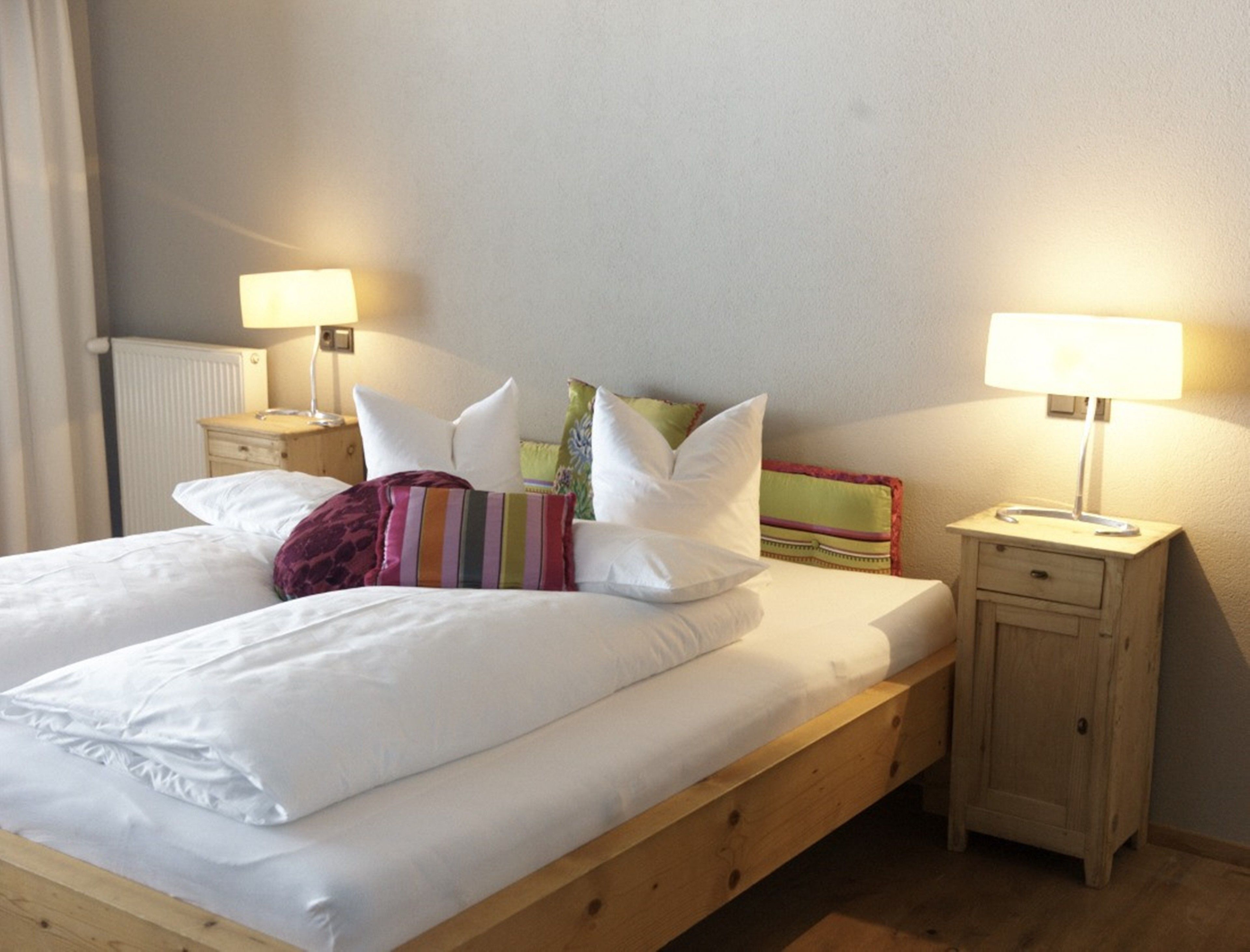 A neatly made bed with white linens and multiple pillows, a wooden nightstand with a lamp, and a radiator on the wall.