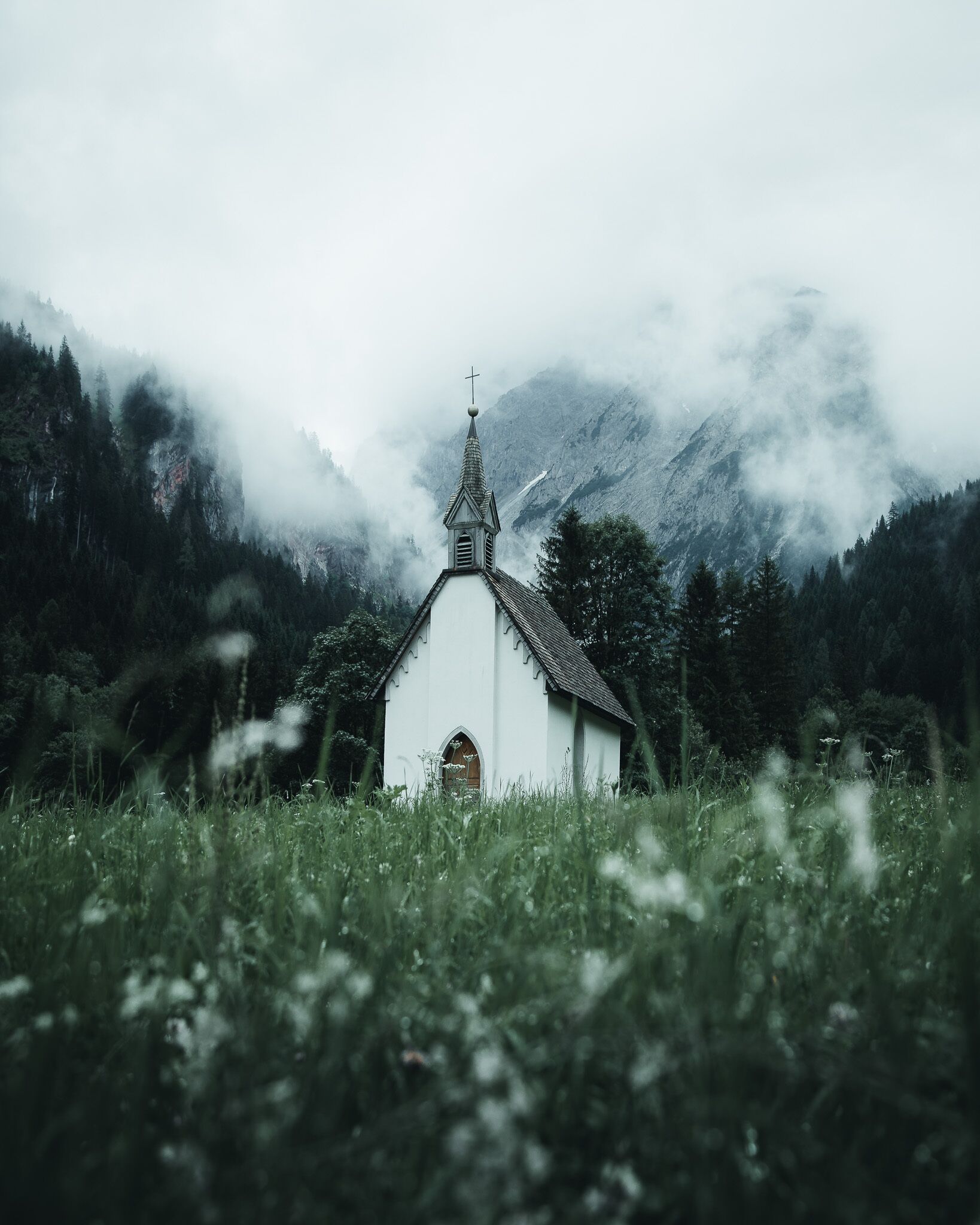 A white church with a pointed steeple and a cross on top, surrounded by a forested mountain backdrop.