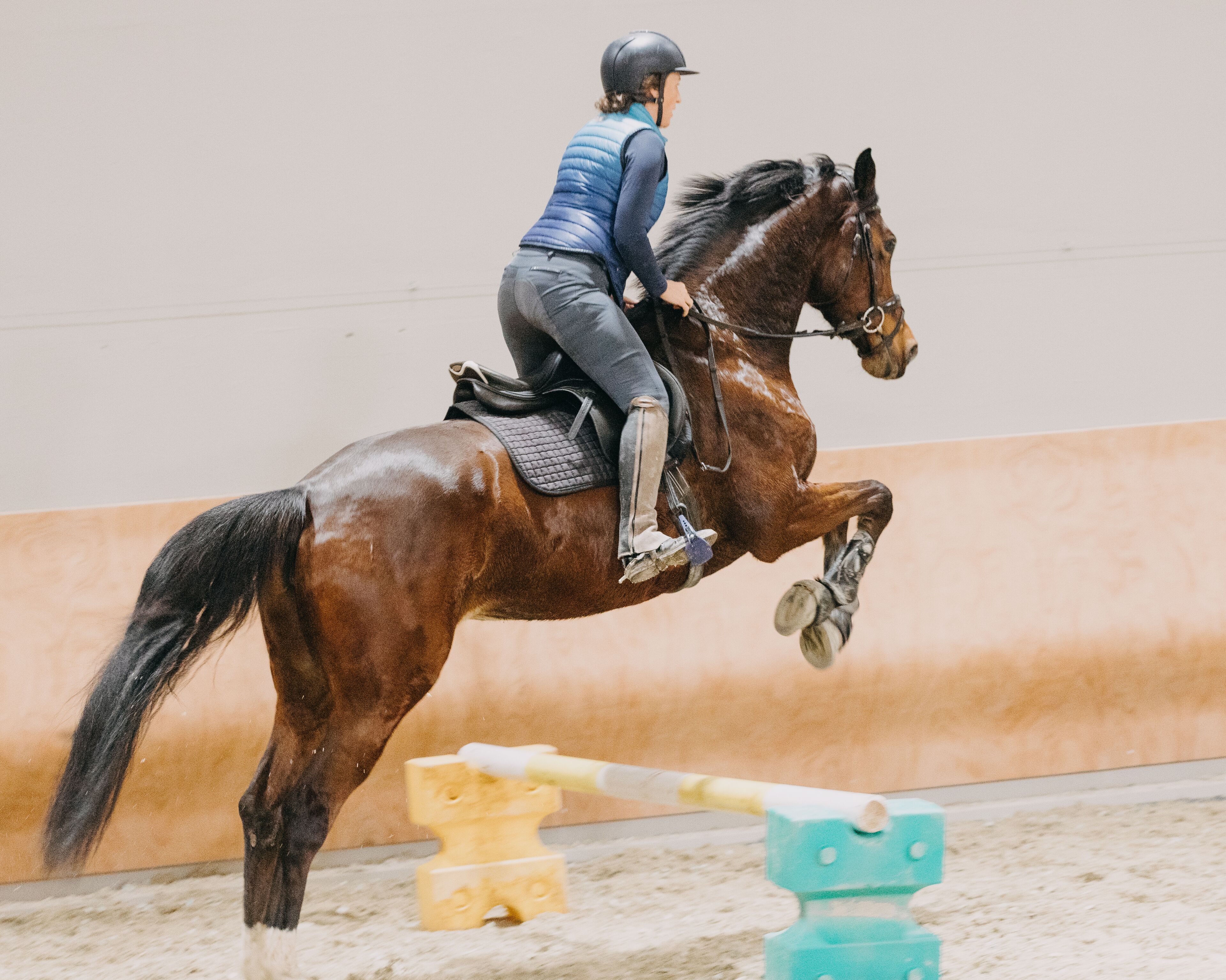 A person riding a horse in an indoor arena with a sandy floor and a padded jump obstacle.