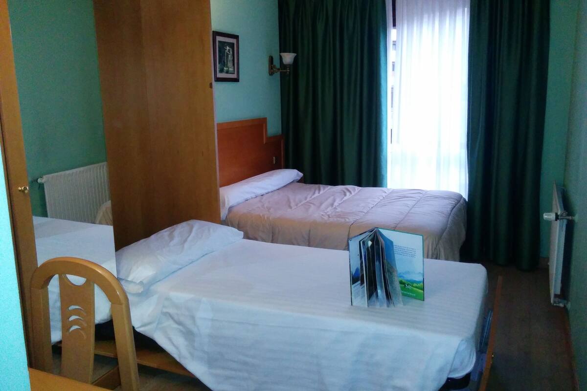 Habitación de hotel con dos camas, una puerta de madera, una silla, un radiador y una ventana con cortinas.