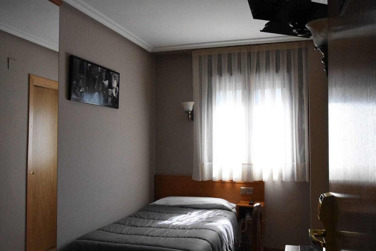 Habitación de hotel con cama, mesita de noche, lámpara, un cuadro en la pared y una ventana con cortinas.