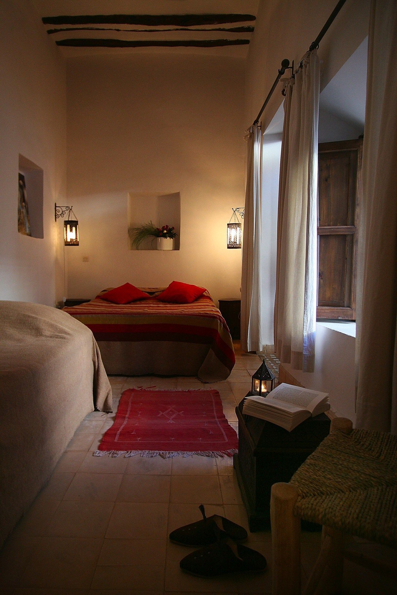 Een gezellige slaapkamer met een bed, een zithoek met een boek en lamp, en een raam met gordijnen.