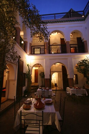 Un área de comedor al aire libre con mesas y sillas, rodeada de arcos y un edificio con balcones.