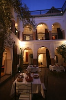 Un área de comedor al aire libre con mesas y sillas, rodeada de arcos y un edificio con balcones.