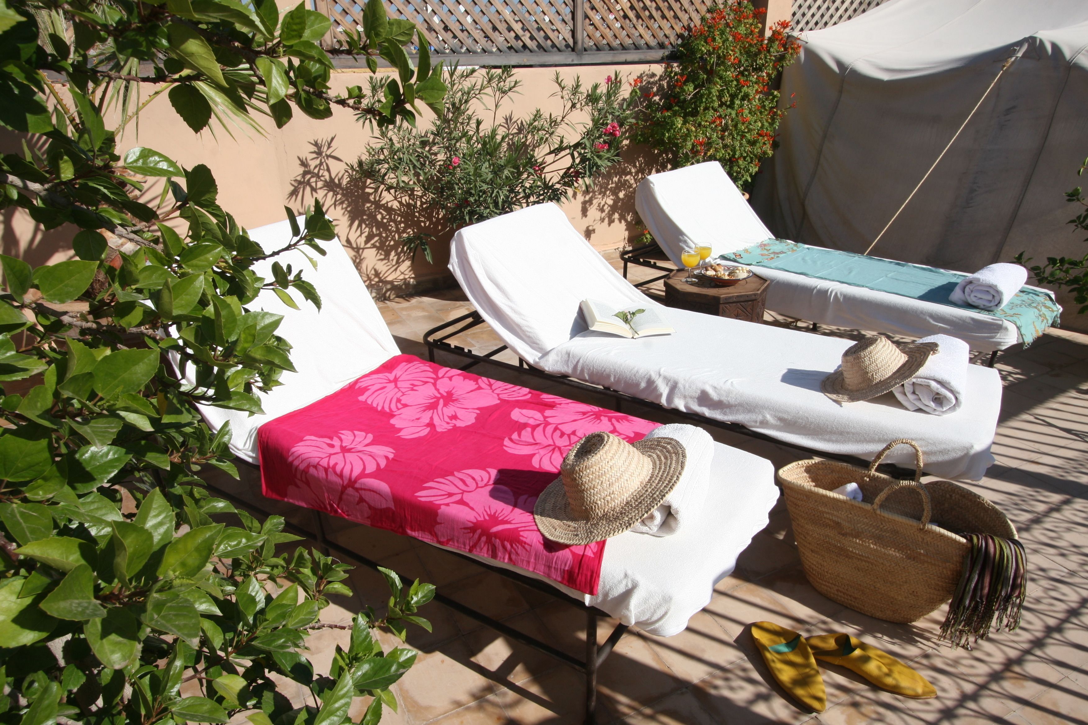 Two sun loungers with towels and a straw hat, a woven basket, and yellow slippers on a patio with a red floral tablecloth.