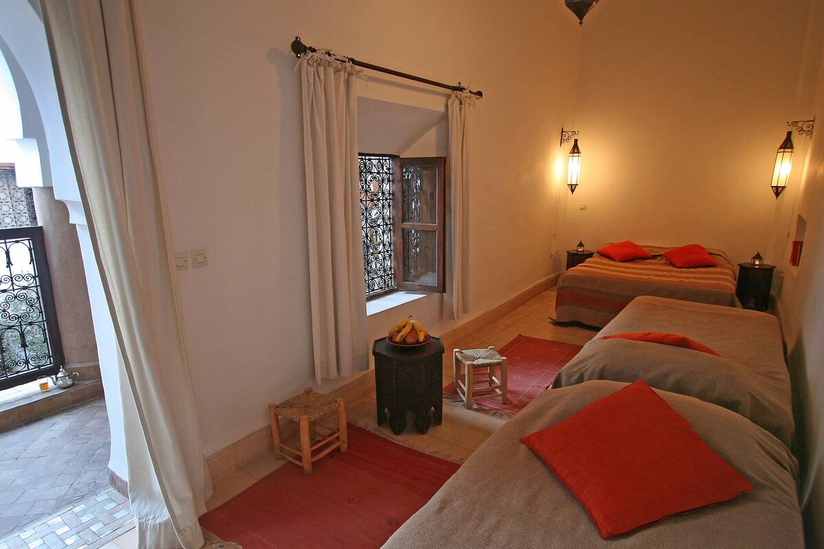 Un dormitorio con dos camas, una ventana con cortinas, un taburete pequeño y una alfombra roja.