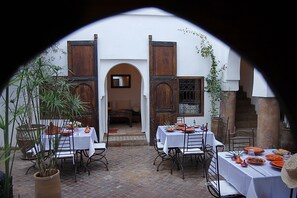 An outdoor dining area with tables set for a meal, surrounded by potted plants and a white building with wooden doors. - Riad Elixir (Marrakech)