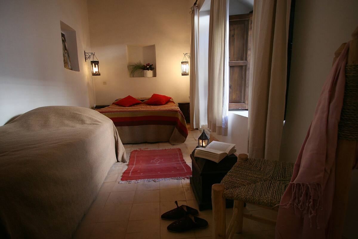 Un dormitorio con cama, una silla, una mesita y una ventana con cortinas.