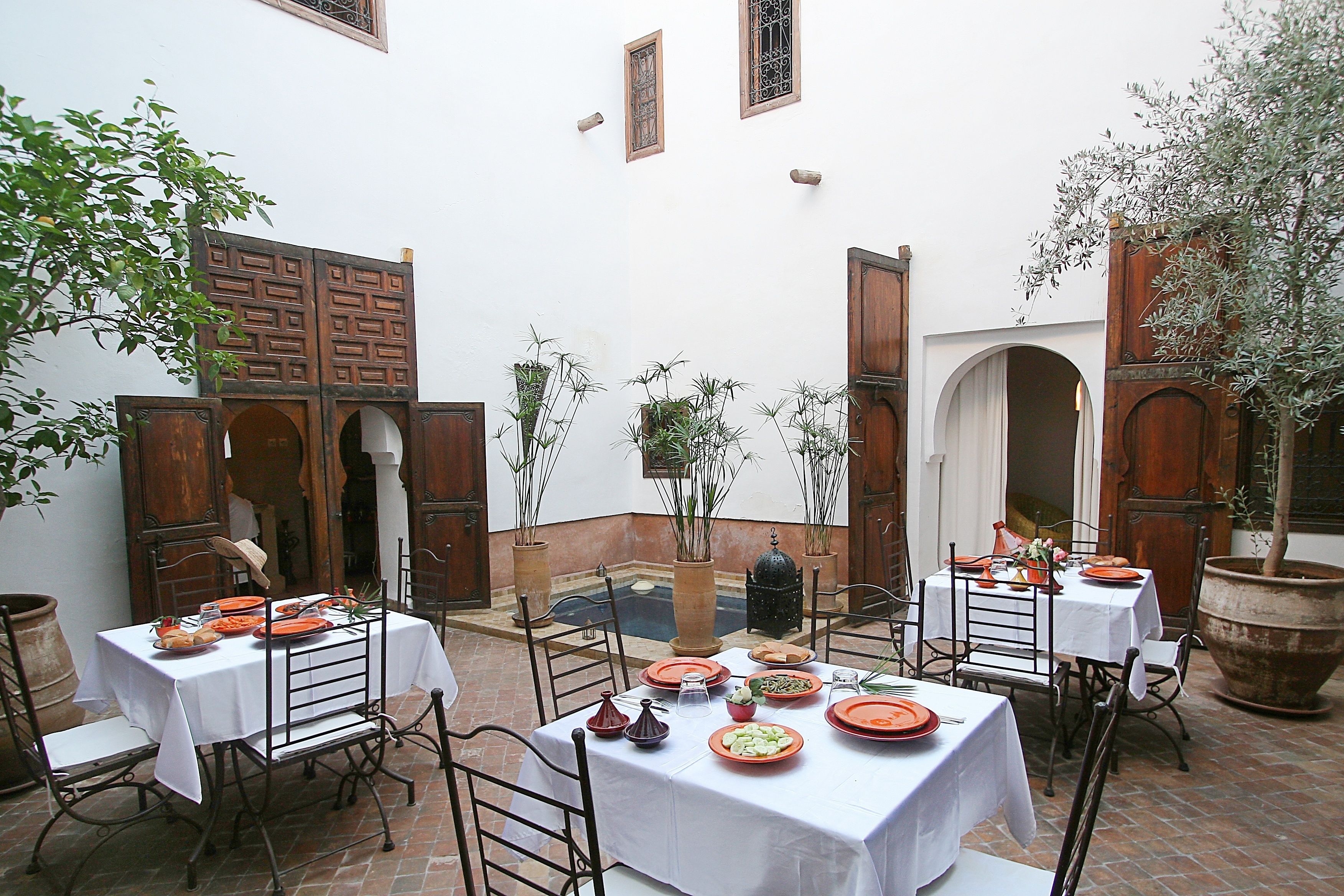Un área de comedor al aire libre con mesas dispuestas para comidas, rodeada de plantas en macetas y detalles arquitectónicos.