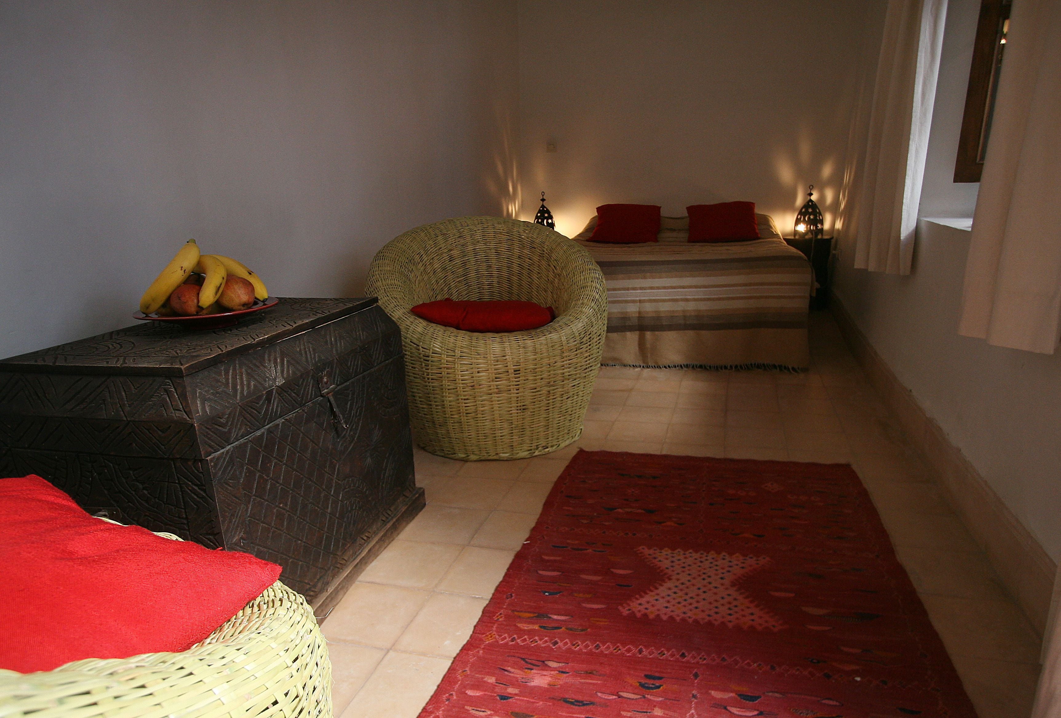 Una camera d'albergo compatta con un letto, una poltrona in vimini, un tavolino con frutta e un tappeto rosso.