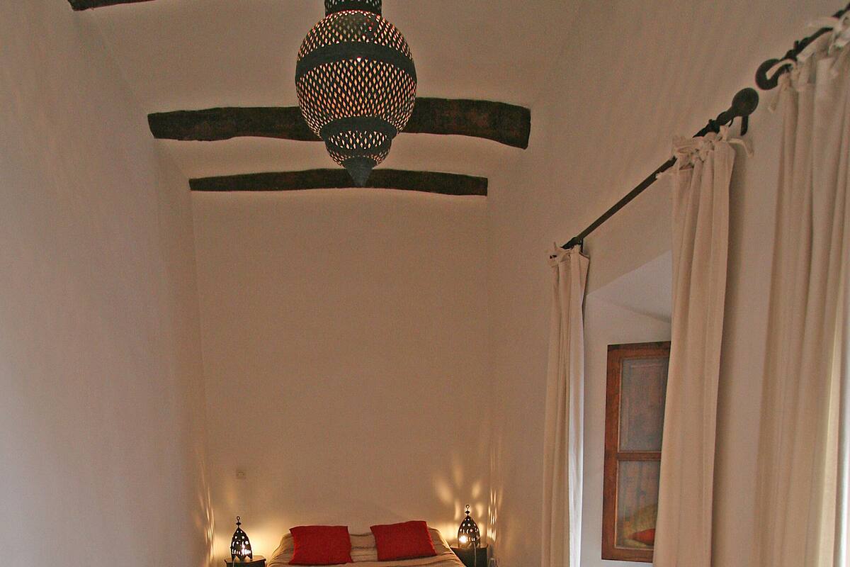 Una habitación pequeña con una cama, una mesita con fruta, una silla de mimbre y una alfombra roja.