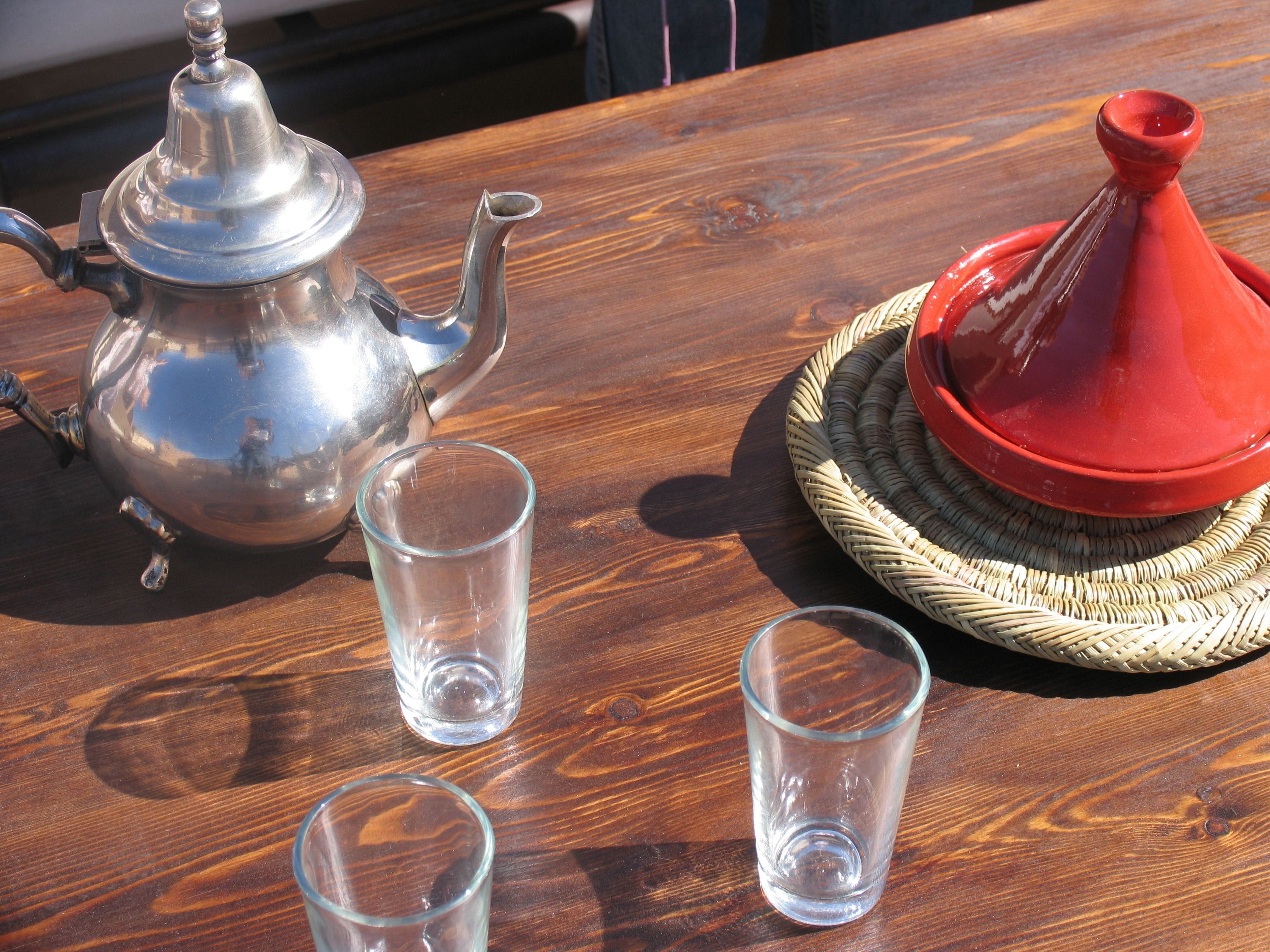 Une table en bois sur laquelle sont posés un théière en argent, deux verres transparents et un tajine rouge et blanc, le tout sur un tapis tissé.
