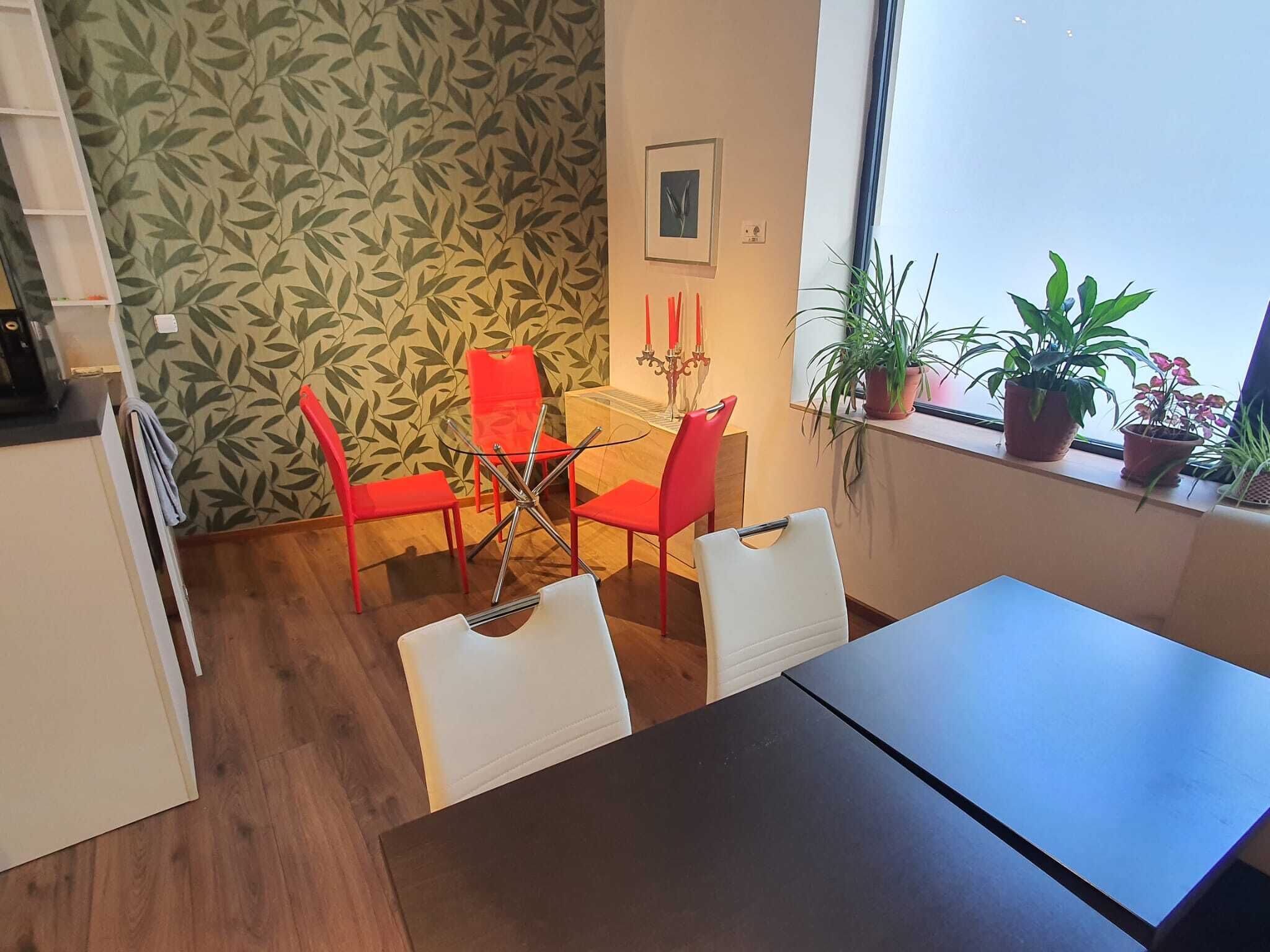 Ein Essbereich mit Tisch und Stühlen, ein Fenster mit Pflanzen und einer Wand mit Blättermuster.