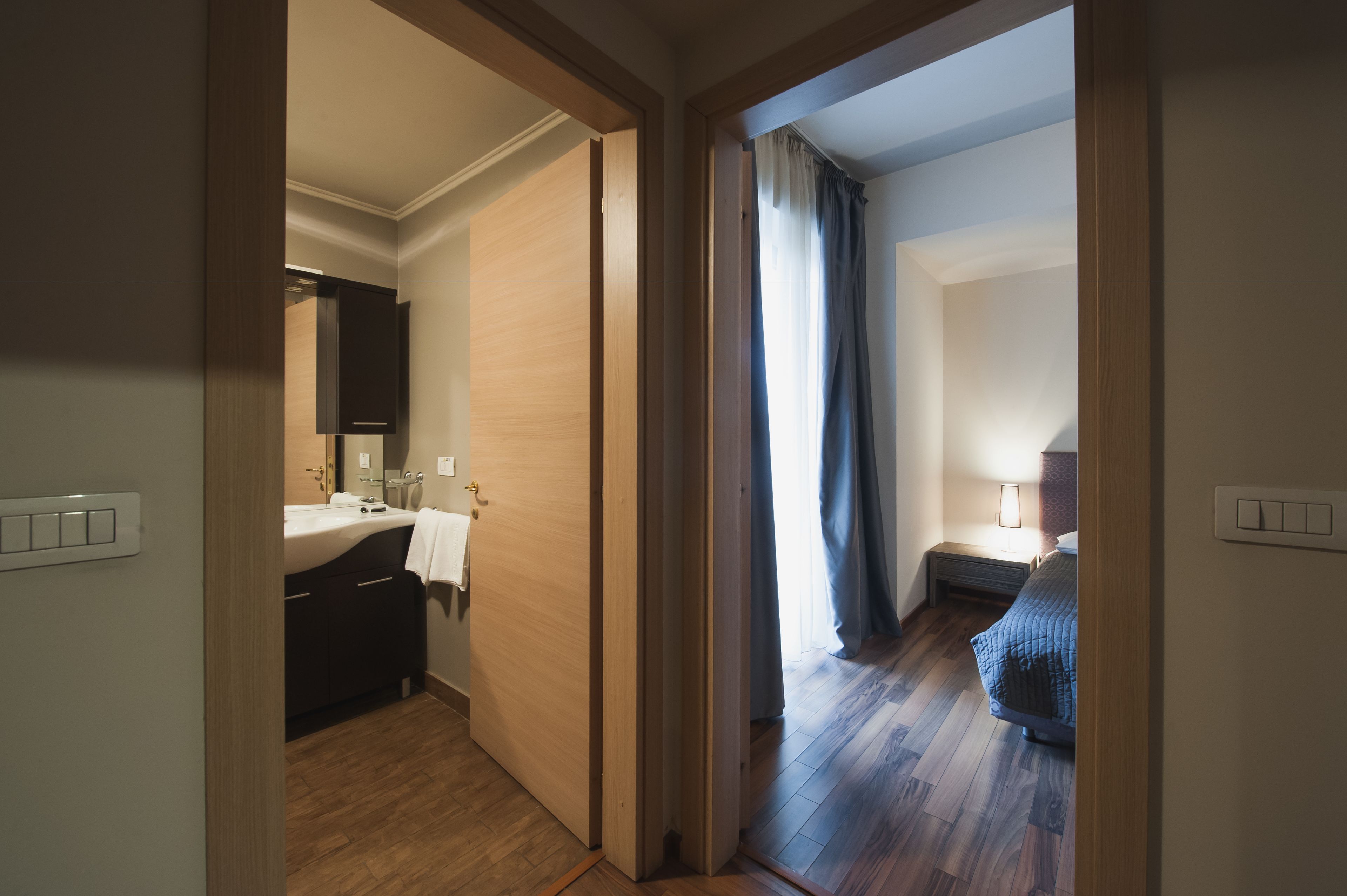 Une chambre d’hôtel avec un plancher en bois, un lit, une table de chevet avec une lampe, une salle de bain avec un lavabo et un miroir, et une porte donnant sur une autre pièce.