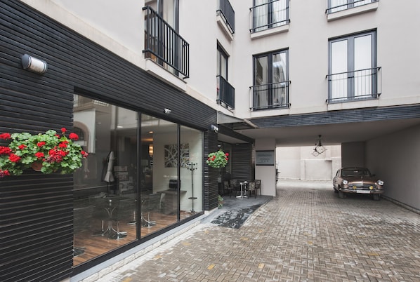 A courtyard with a car parked in a covered area, a building with a glass facade, and a hanging flower arrangement. - Hemingway Residence (Bucharest)