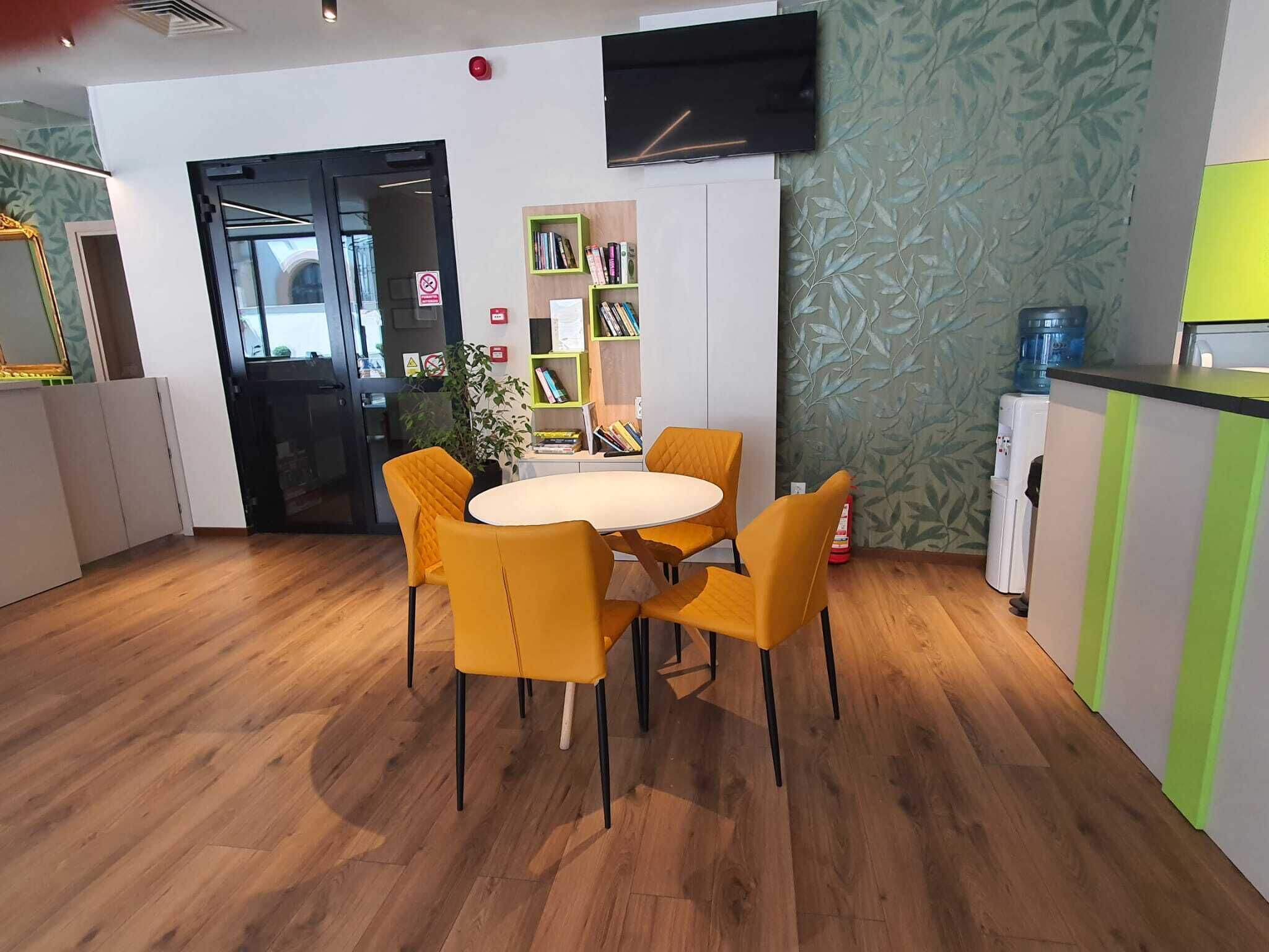 Uma sala de jantar moderna com uma mesa redonda e quatro cadeiras amarelas, uma estante, um dispensador de água e uma televisão de tela plana na parede.