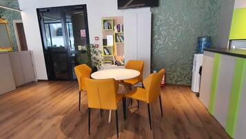 Uma sala de jantar moderna com uma mesa redonda e quatro cadeiras amarelas, uma estante, um dispensador de água e uma televisão de tela plana na parede.