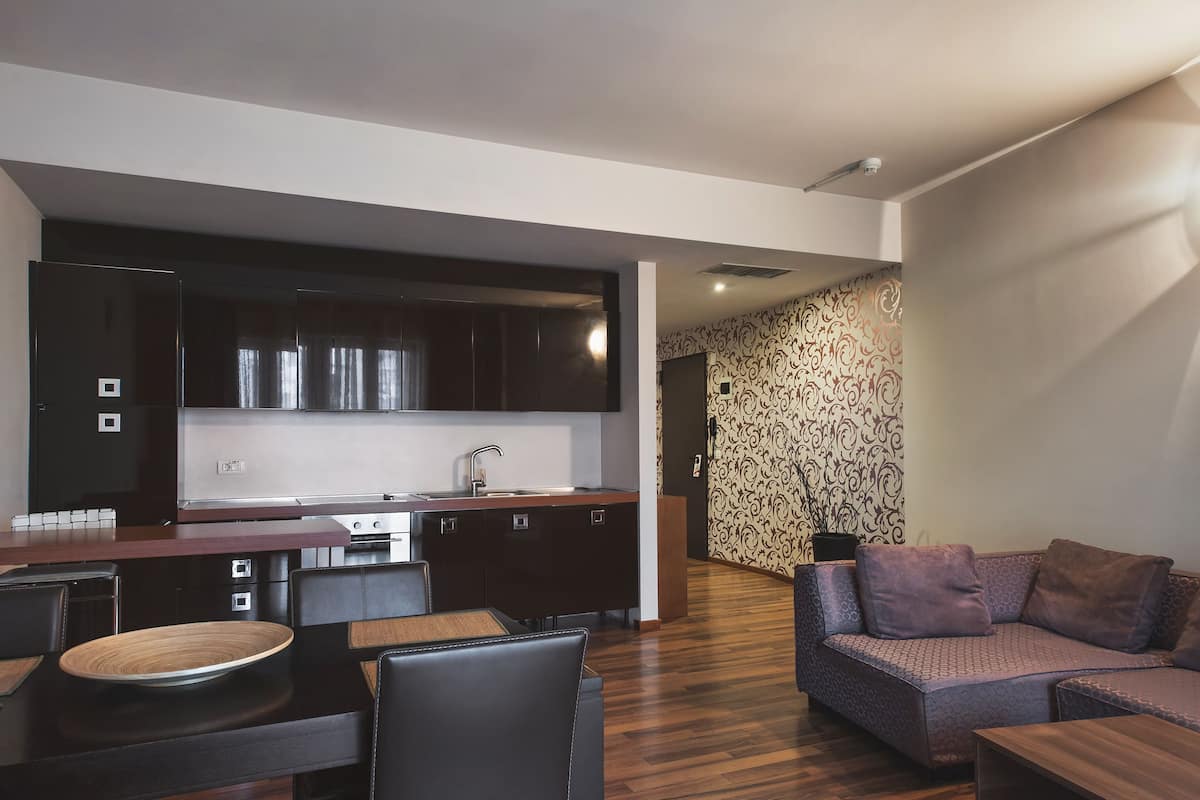Eine moderne Küche mit dunklen Schränken und einer Edelstahl-Dunstabzugshaube, ein Essbereich mit Holztisch und -stühlen sowie ein Wohnzimmer mit einem gemusterten Sofa und einem Couchtisch aus Holz.