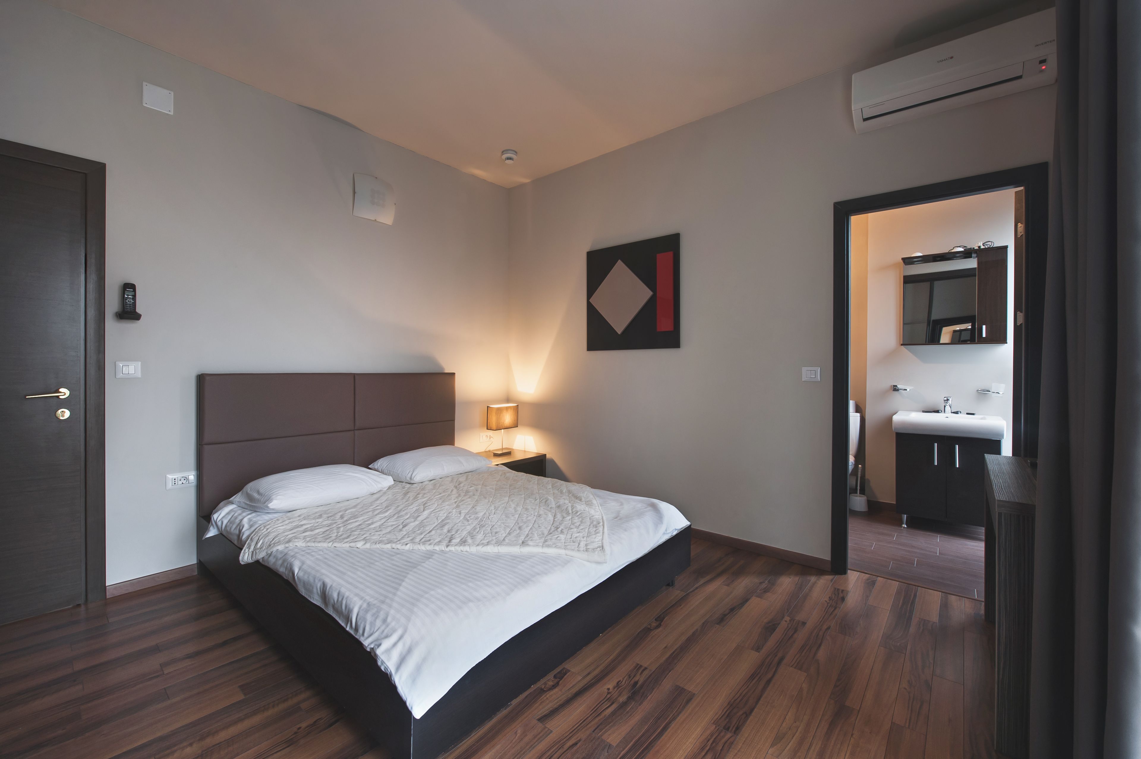 Une chambre à coucher avec un lit, une table de chevet, une lampe, et une salle de bain visible à travers une porte ouverte.