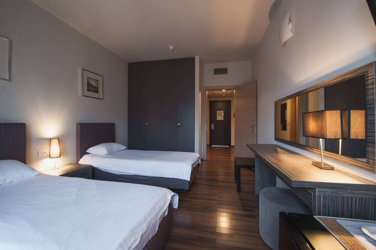 Ein Hotelzimmer mit zwei Betten, einem Schreibtisch, einem Spiegel und einer Tür zu einem weiteren Raum.