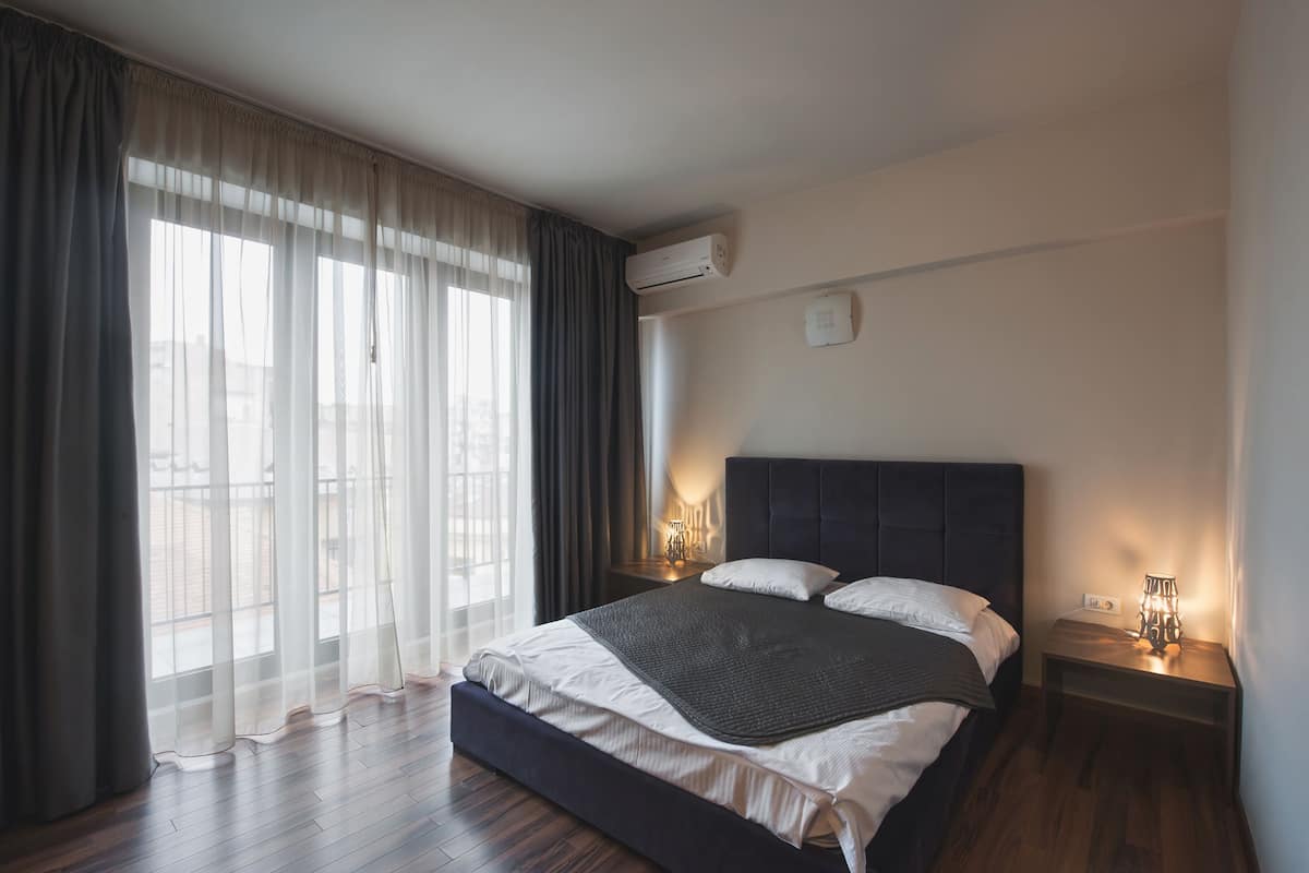 Ein Schlafzimmer mit einem großen Bett, einem Nachttisch mit Lampe und einem Fenster mit durchsichtigen Vorhängen.
