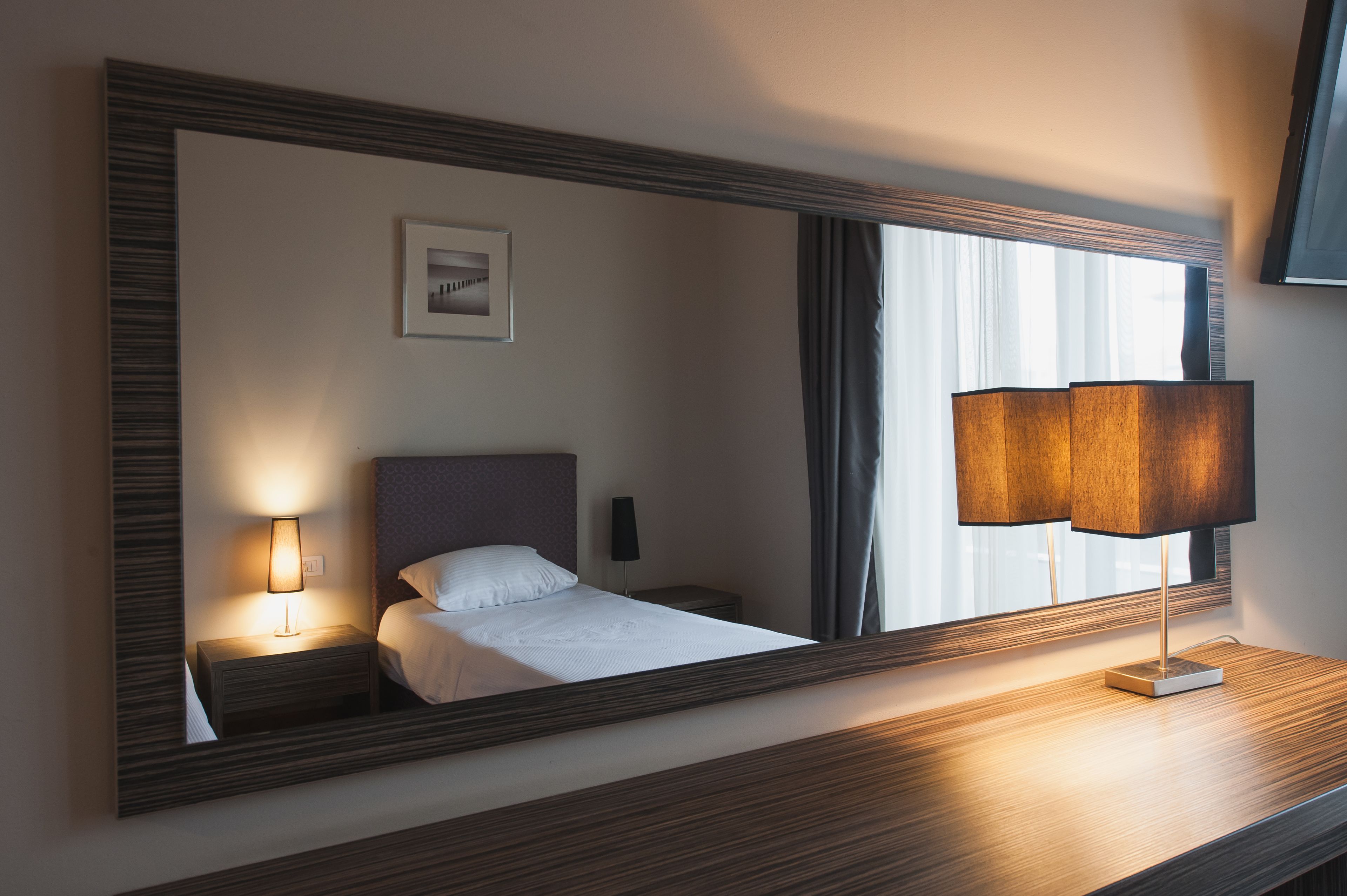 A hotel room with a large mirror, a bed, a bedside lamp, a nightstand, and a window with curtains.