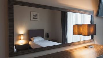 A hotel room with a large mirror, a bed, a bedside lamp, a nightstand, and a window with curtains.