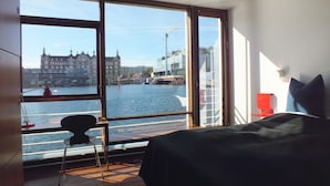 A room with a large window overlooking a river, a bed, a chair, and a red chair. - CPH Living (Copenhagen)