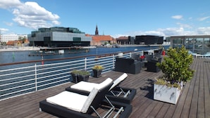 A deck with lounge chairs and planters, overlooking a river with buildings in the background. - CPH Living (Copenhagen)