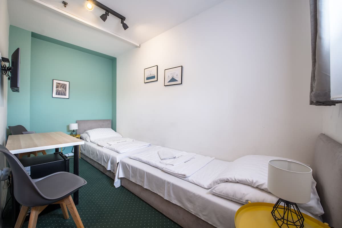 A small, neatly arranged room with a bed, a desk, and a chair. There are two framed pictures on the wall, a lamp on the desk, and a yellow tray on the bed.