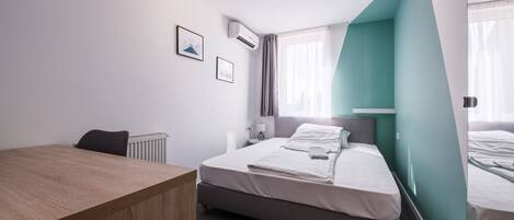 Una camera d'albergo moderna con una scrivania in legno, un letto con lenzuola bianche, un poggiatesta grigio e una parete verde acqua.