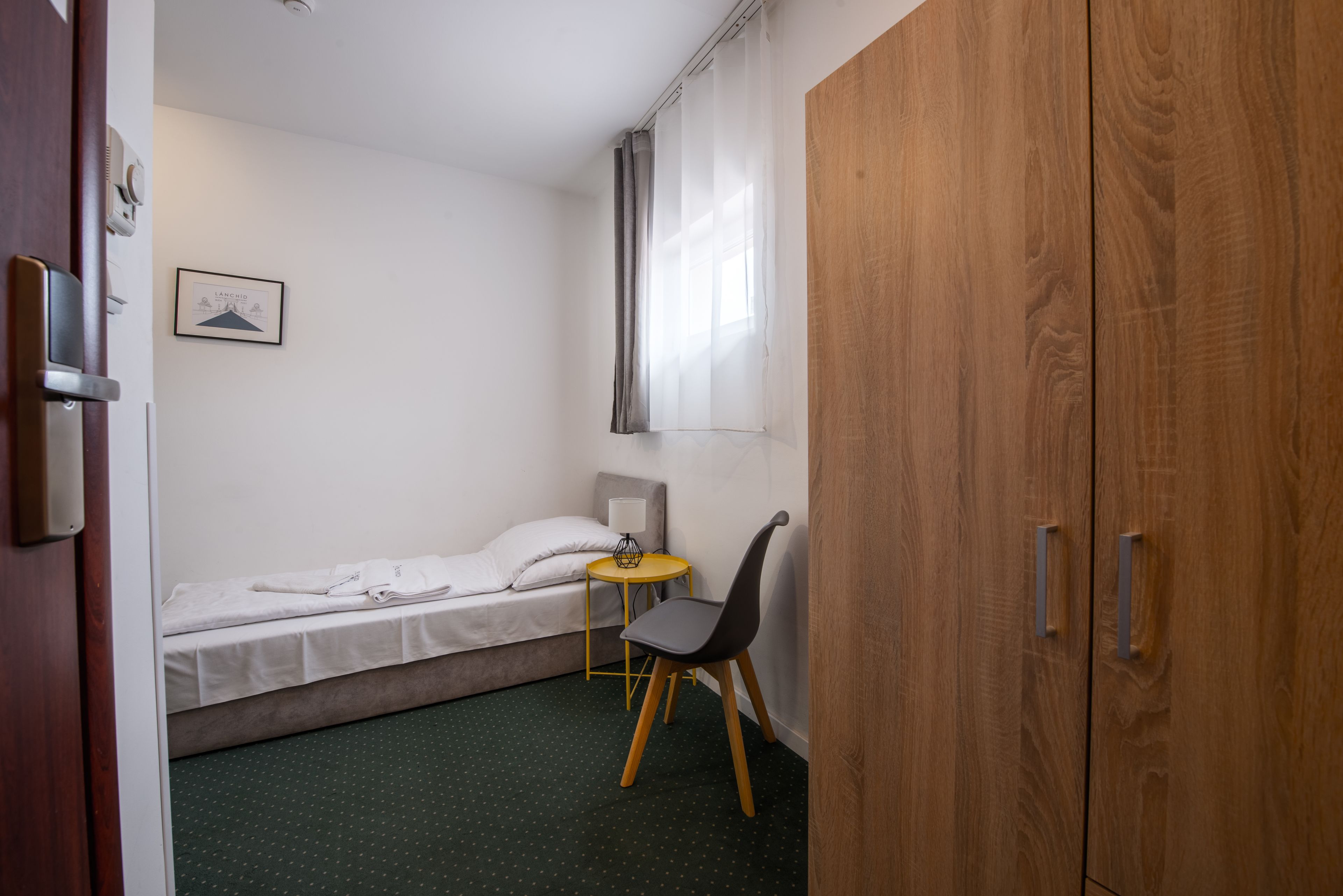 A small, neatly arranged room with a bed, a chair, a small table, and a wardrobe.