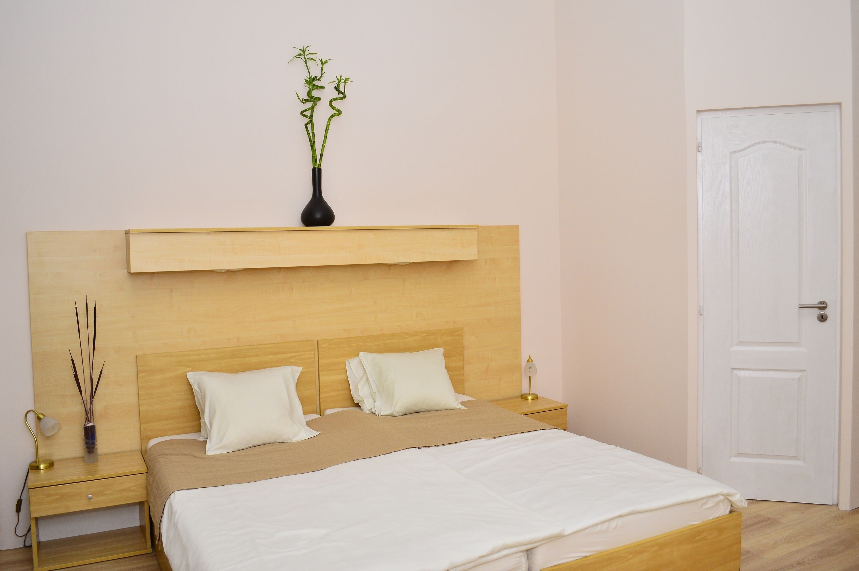Ein ordentlich eingerichtetes Schlafzimmer mit einem Holzkopfteil, zwei weißen Kissen, einer beigen Bettdecke, einem Nachttisch mit Lampe und einer Vase mit Grünpflanzen.