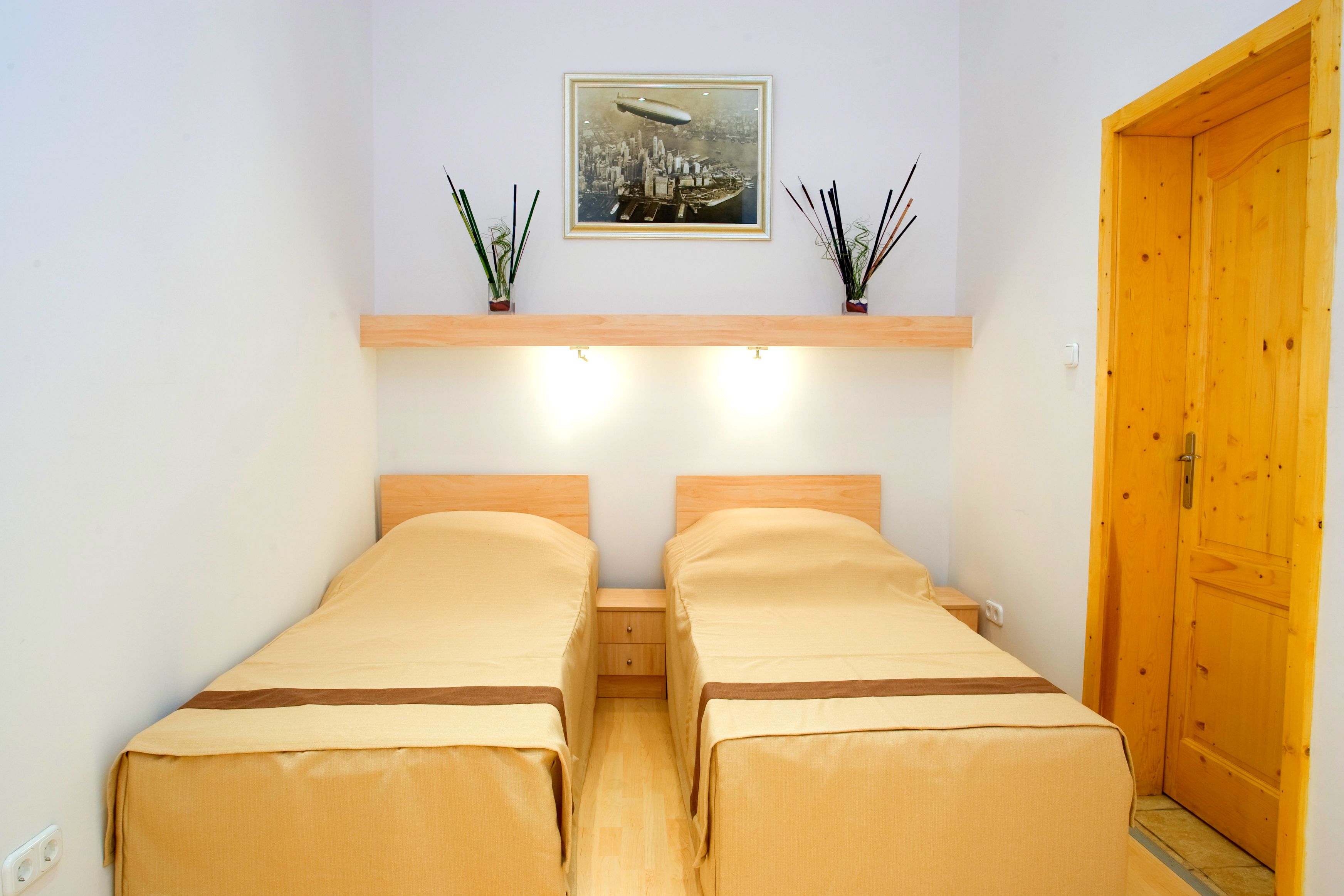Une chambre avec deux lits simples, une porte en bois, une table de chevet, un tableau encadré au mur et une étagère avec des objets décoratifs.