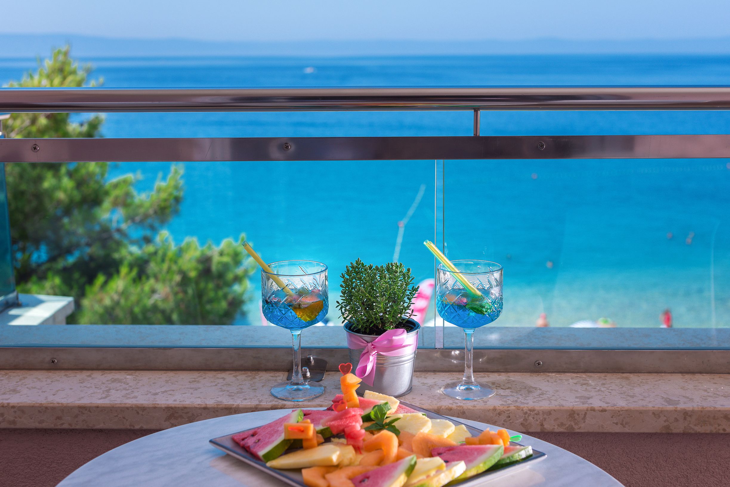 A balcony with a view of the sea, two drinks with straws, a small potted plant, and a plate of fruit.