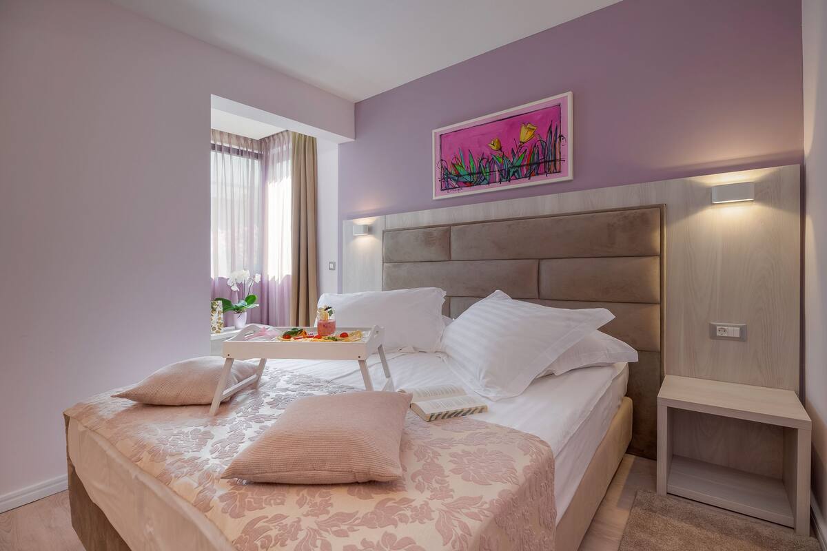 A bedroom with a bed, bedside tables, a nightstand, a window with curtains, and a wall-mounted light fixture.