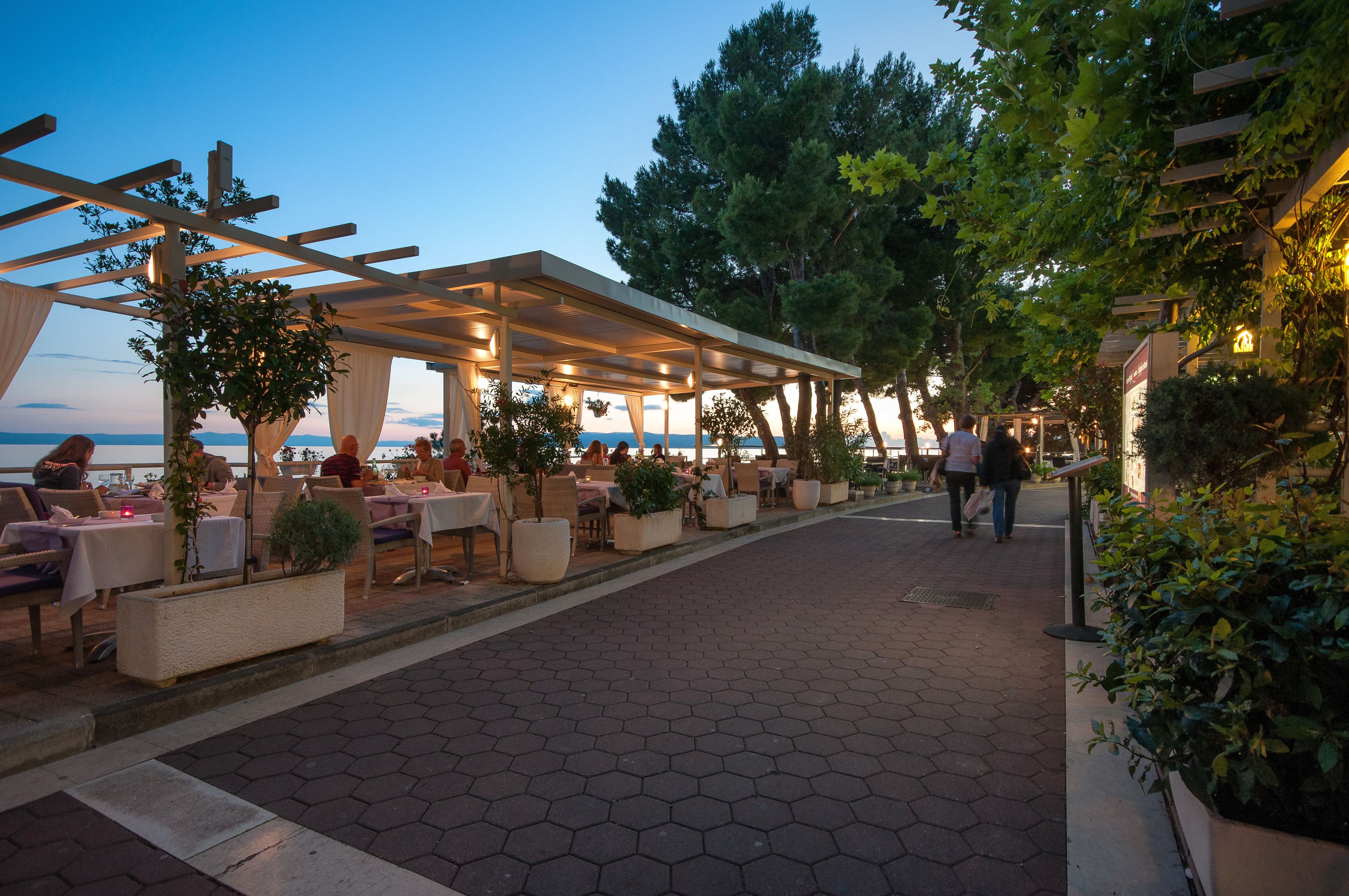 An outdoor dining area with tables and chairs, a covered walkway, and a view of the sea.